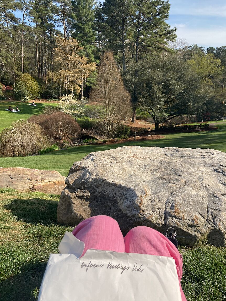 in einer rosa Hose gekleidete Knie vor einem schön angelegten Park; auf den Knien ein Umschlag mit den handgeschriebenen Worten: "Conference Readings Duke"