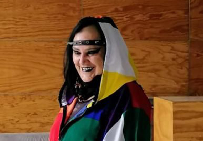 portrait of María Galindo wearing a colourful hoodie, a black studded head band and black lipstick